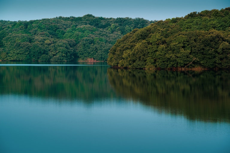 A calm lake reflects dense green trees along the shoreline, with a clear, blue sky above. The water is still, creating a mirror-like image of the forested landscape.