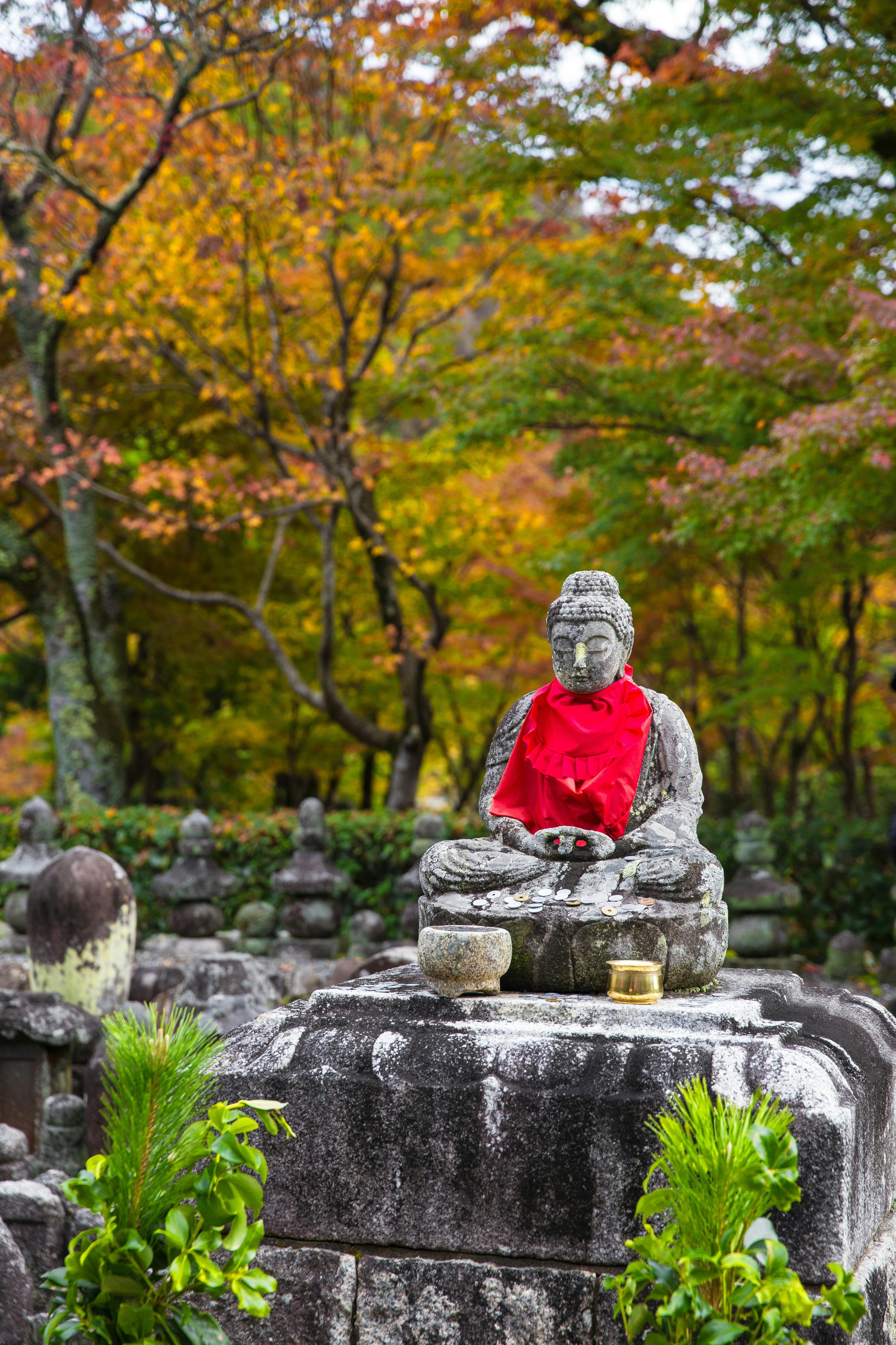 A stone Buddha statue draped in a red bib is seated on a pedestal surrounded by small offerings, with vibrant autumn trees in the background.