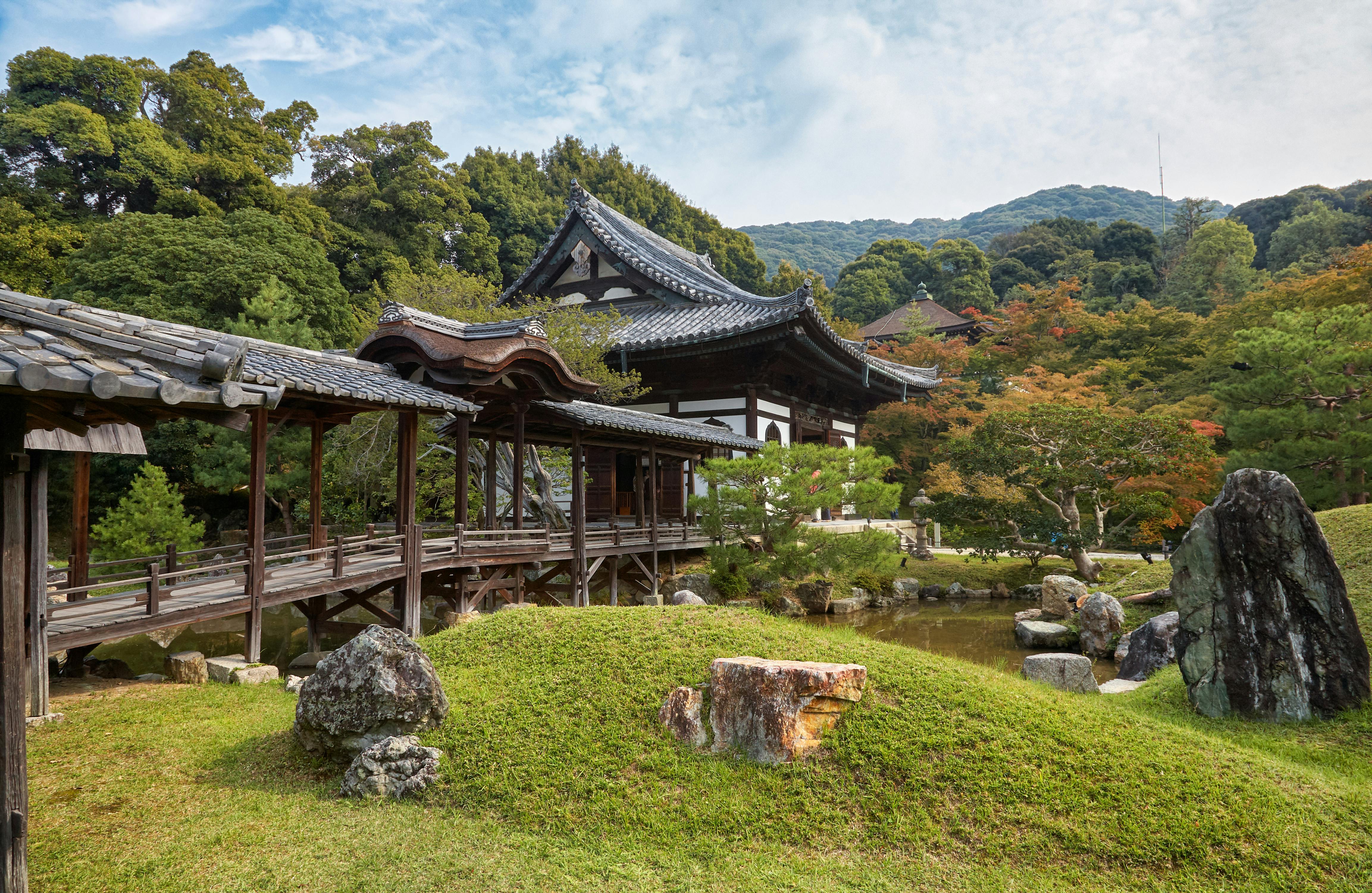 A traditional Japanese building with a covered wooden walkway sits amid lush greenery, manicured grass, rocks, and trees, with forested hills rising in the background under a partly cloudy sky.