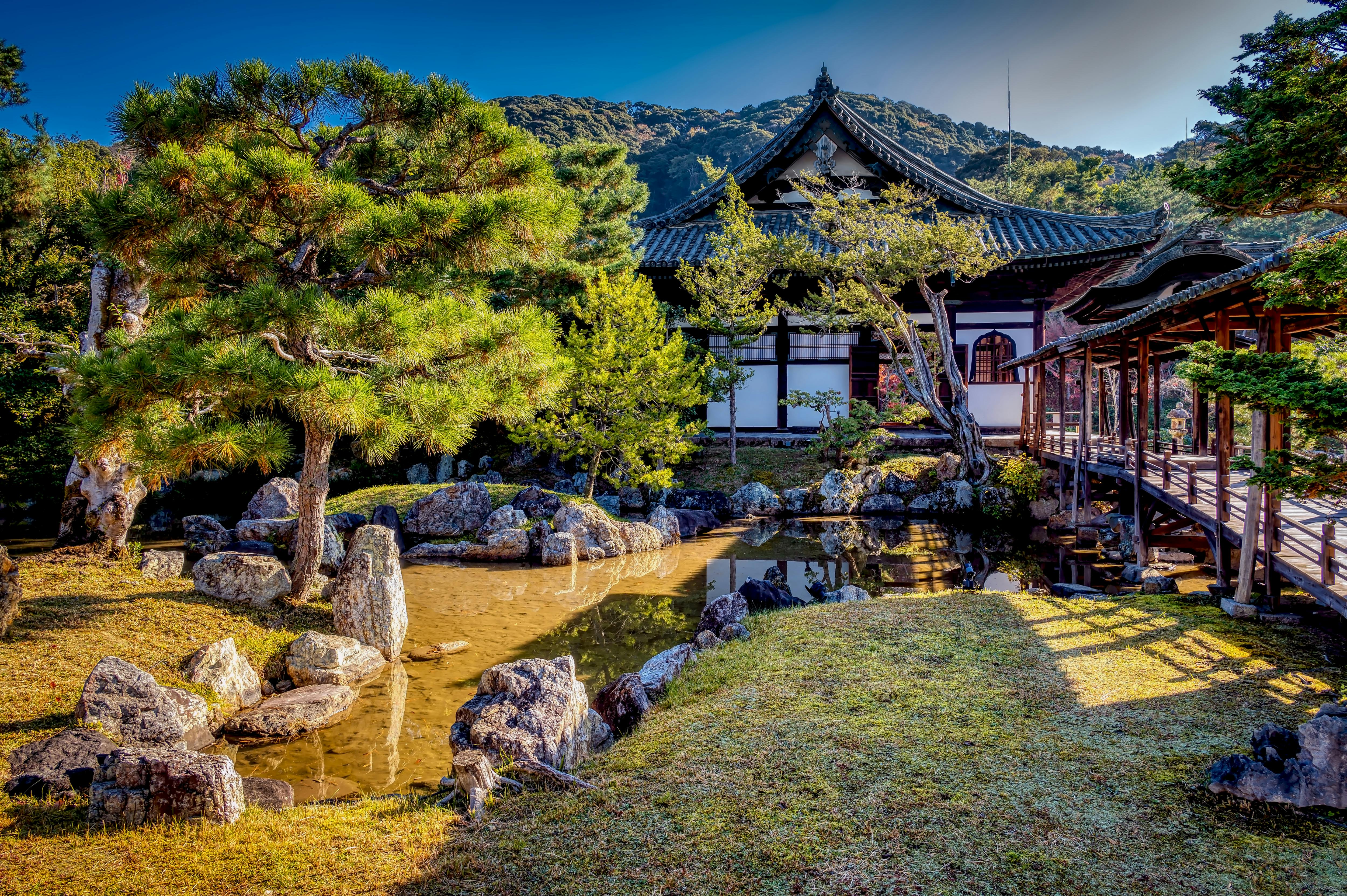 Traditional Japanese garden with a pond, rocks, and manicured trees beside a historic building and wooden walkway, set against a backdrop of forested hills under a clear blue sky.
