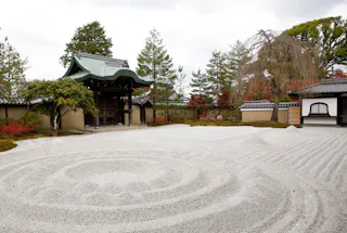 A traditional Japanese Zen garden with carefully raked white gravel forming circular and linear patterns, surrounded by trees, a wooden gate, and a building with curved roof tiles.