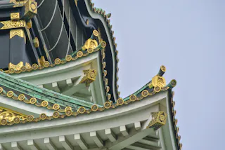 Close-up of the ornate, white and green roof of a traditional Japanese castle with decorative gold accents and intricate architectural details against a clear blue sky.