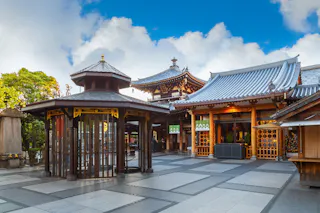 A traditional Japanese temple with tiled roofs, wooden structures, and ornate details, set against a blue sky with scattered clouds. The courtyard is spacious and surrounded by greenery.