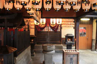 A traditional Japanese shrine interior with hanging lanterns, a stone basin in the center, wooden plaques on the wall, and small altars with incense burners. Warm lighting creates a serene, spiritual atmosphere.