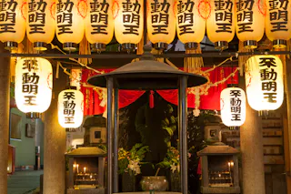 A close-up of a Japanese shrine at dusk, decorated with glowing paper lanterns featuring black kanji characters, a central incense burner, flowers, and candles illuminating the scene.