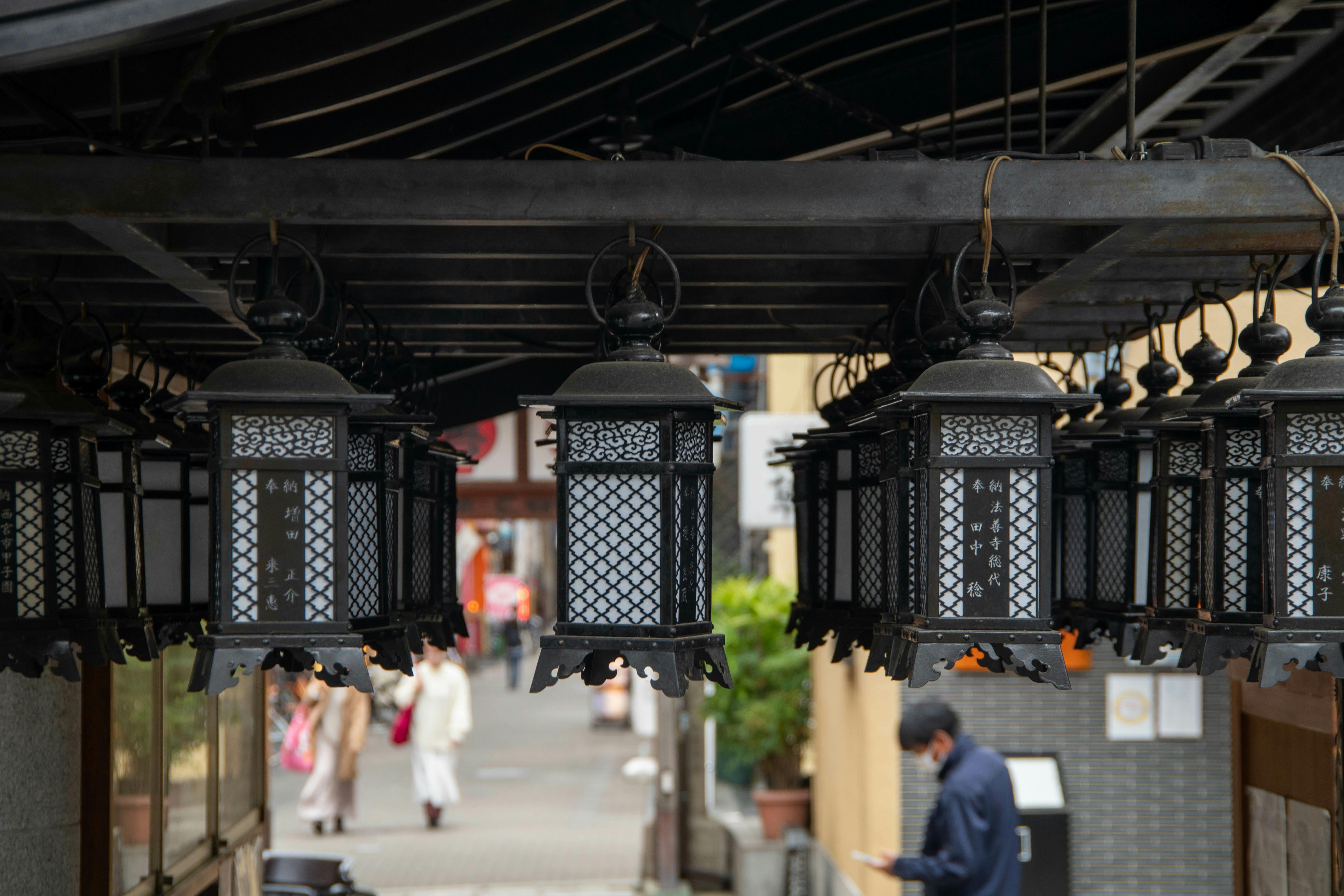 Hanging black metal lanterns under a roof, with a blurred street scene in the background where people are walking and one person appears to be reading or looking at a phone.