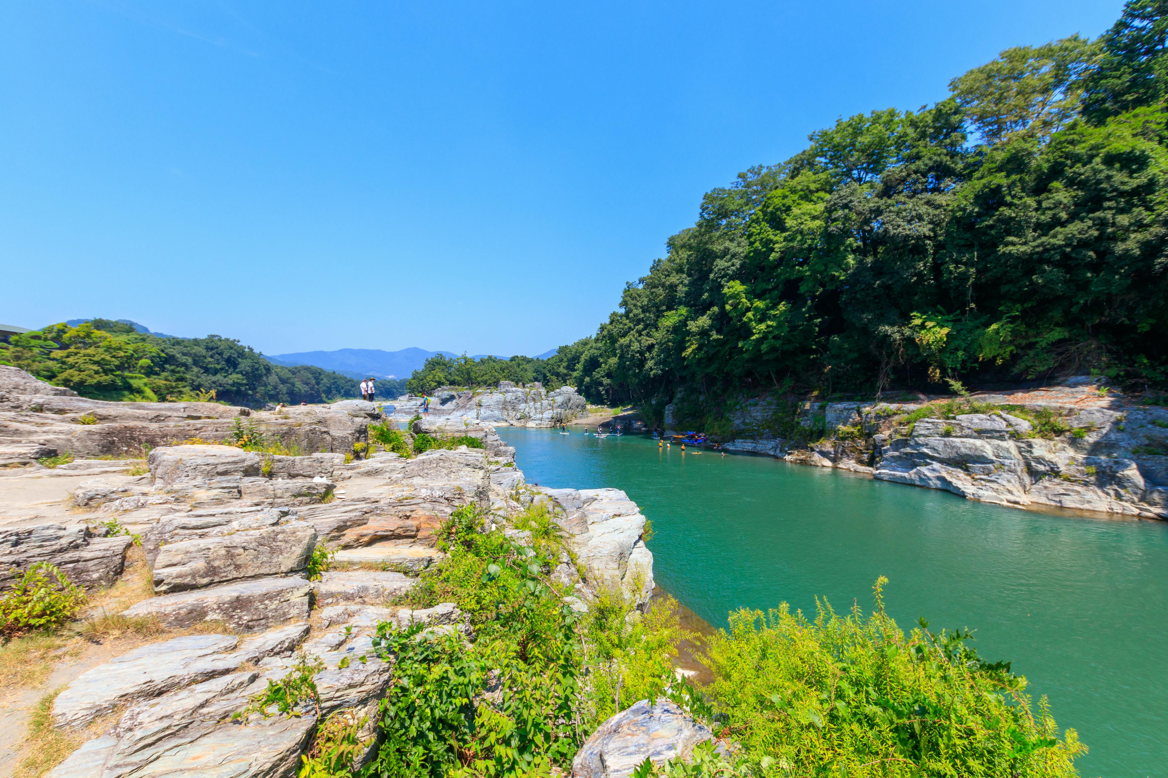 A clear river flows between rocky cliffs and lush green trees under a bright blue sky. Small groups of people stand on the rocks and swim in the water, surrounded by natural scenery.