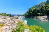 A clear river flows between rocky cliffs and lush green trees under a bright blue sky. Small groups of people stand on the rocks and swim in the water, surrounded by natural scenery.