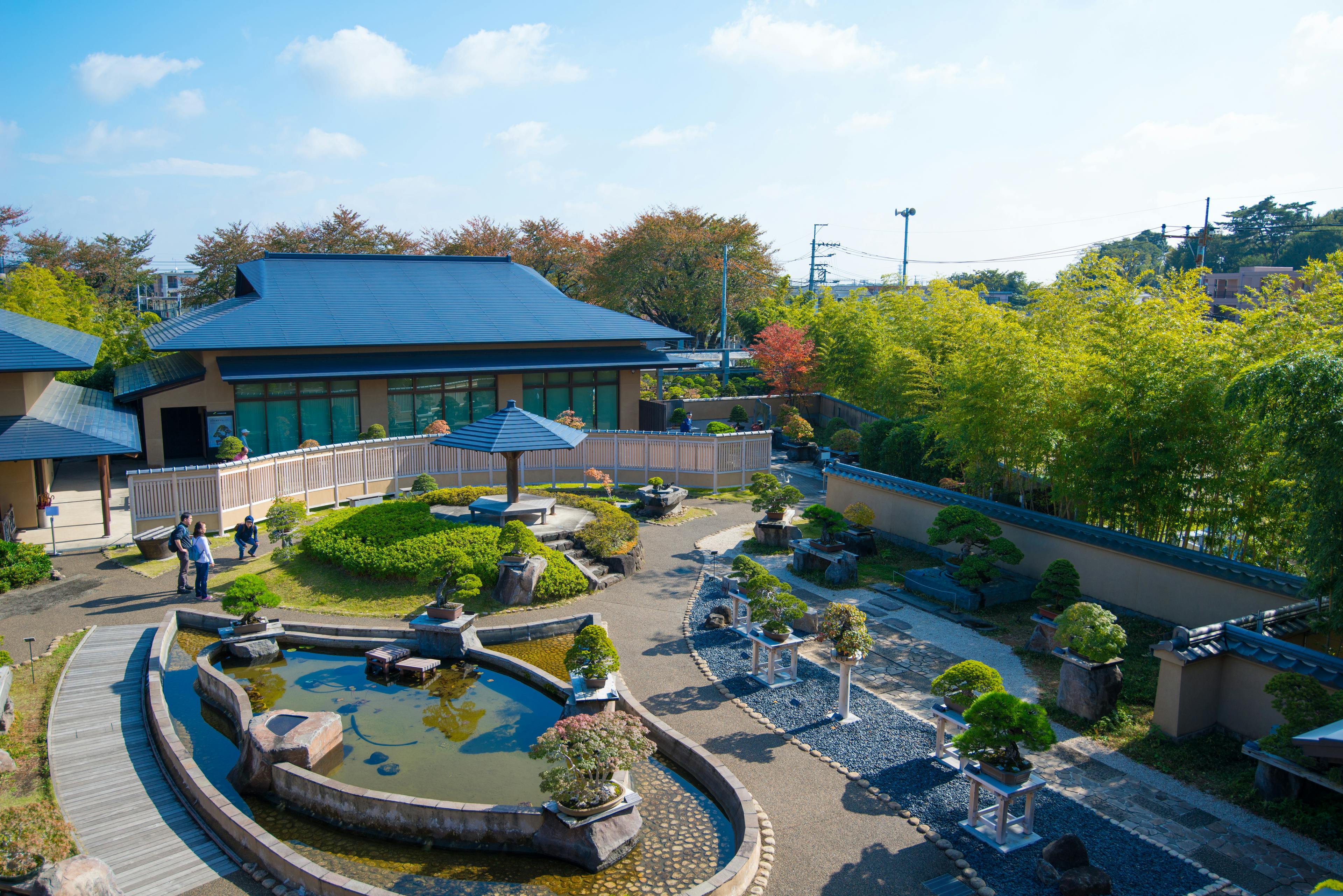 A serene Japanese garden with a pond, manicured bushes, stone paths, and traditional-style buildings surrounded by trees and bamboo under a bright blue sky. A few people are walking along the paths.