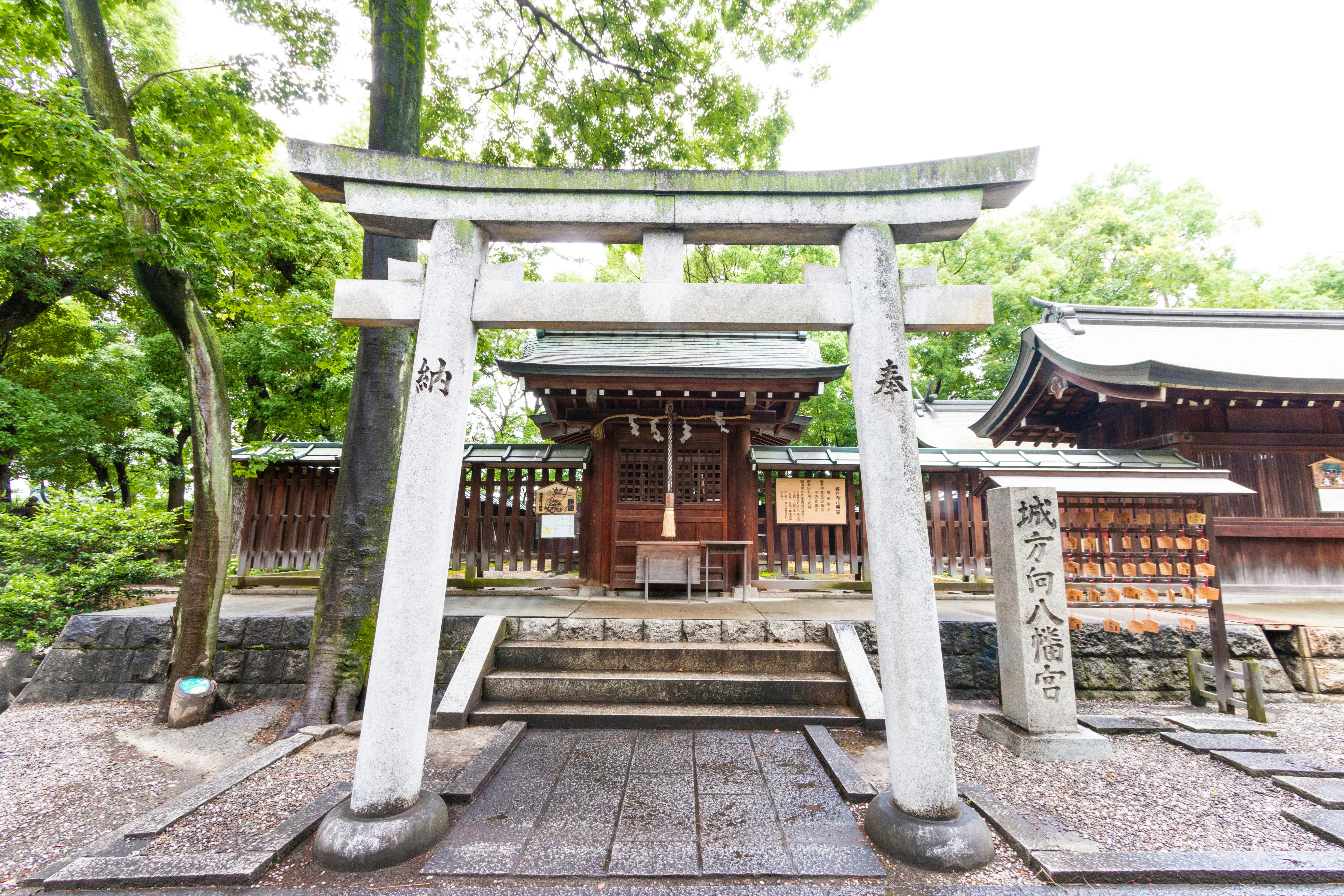 A stone torii gate stands at the entrance of a traditional Japanese Shinto shrine, surrounded by trees and wooden structures, with a stone tablet inscribed with Japanese characters nearby.