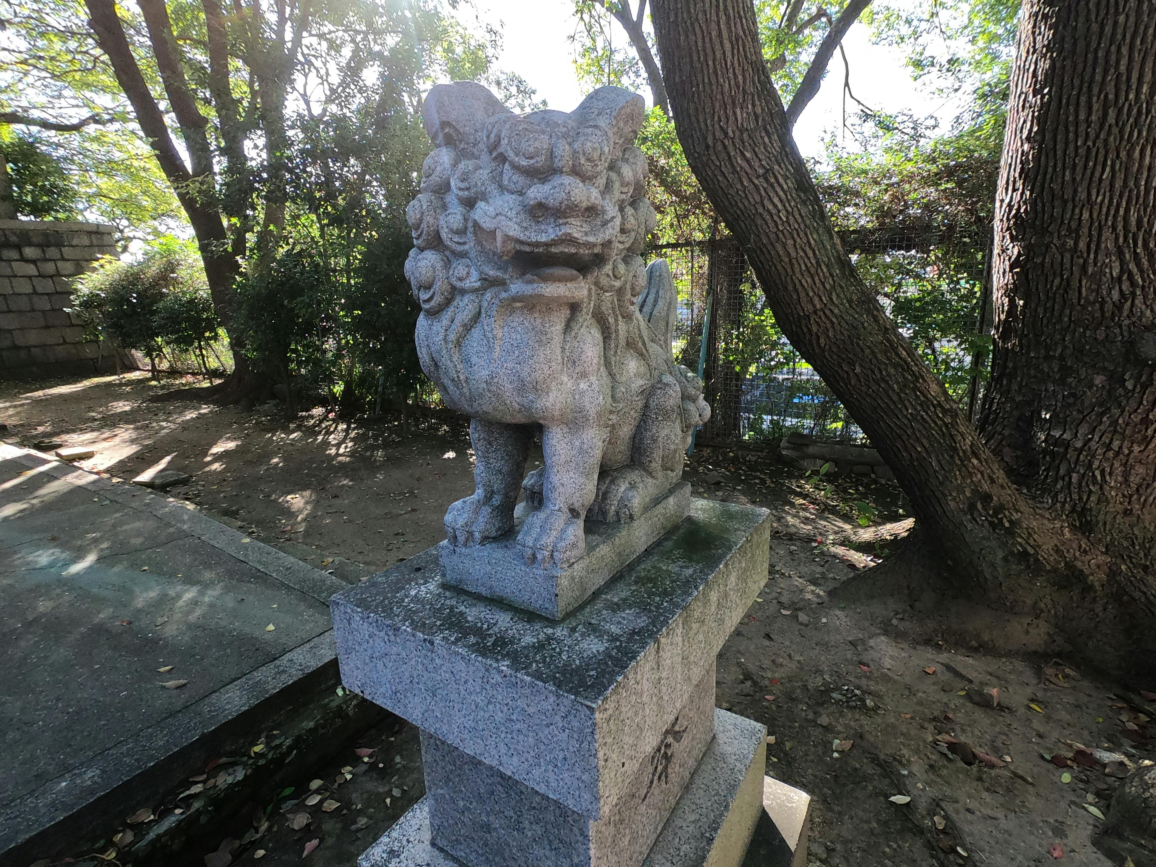 A stone guardian lion-dog statue, known as a komainu, is positioned on a pedestal in a shaded outdoor area surrounded by trees and sunlight filtering through the leaves.