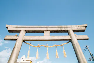 A traditional Japanese torii gate made of stone stands under a clear blue sky, decorated with a rope and straw tassels. Buildings and power lines are visible in the background.