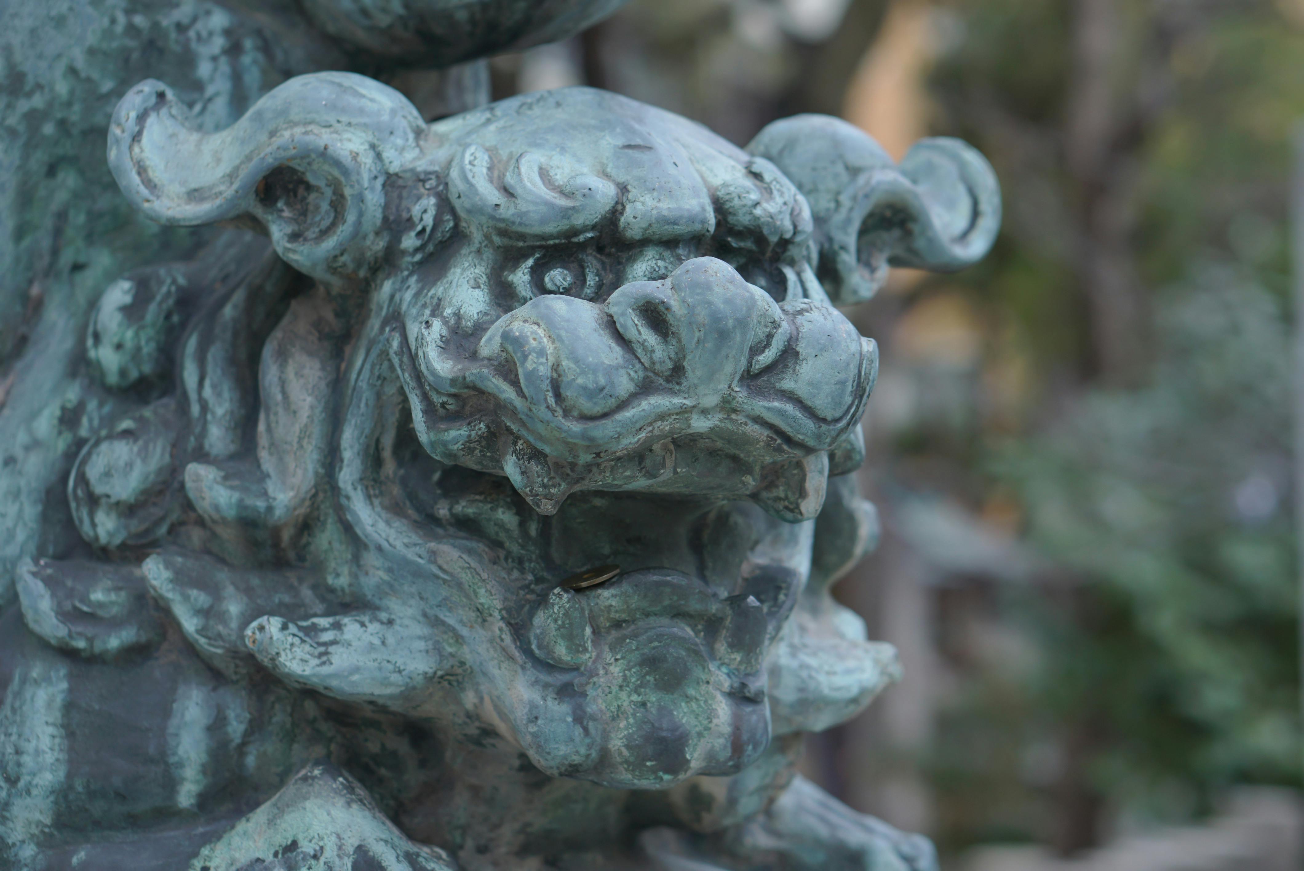 Close-up of a weathered, greenish-blue stone or metal statue of a mythical lion-like creature with an open mouth, showing sharp teeth and fierce expression. The background is blurred trees and outdoor scenery.