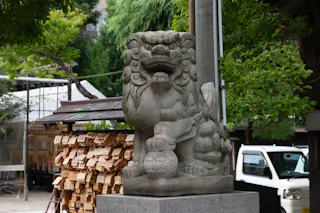 A stone lion-dog statue sits on a pedestal outdoors near a pile of wooden plaques and a white vehicle, surrounded by trees and greenery.