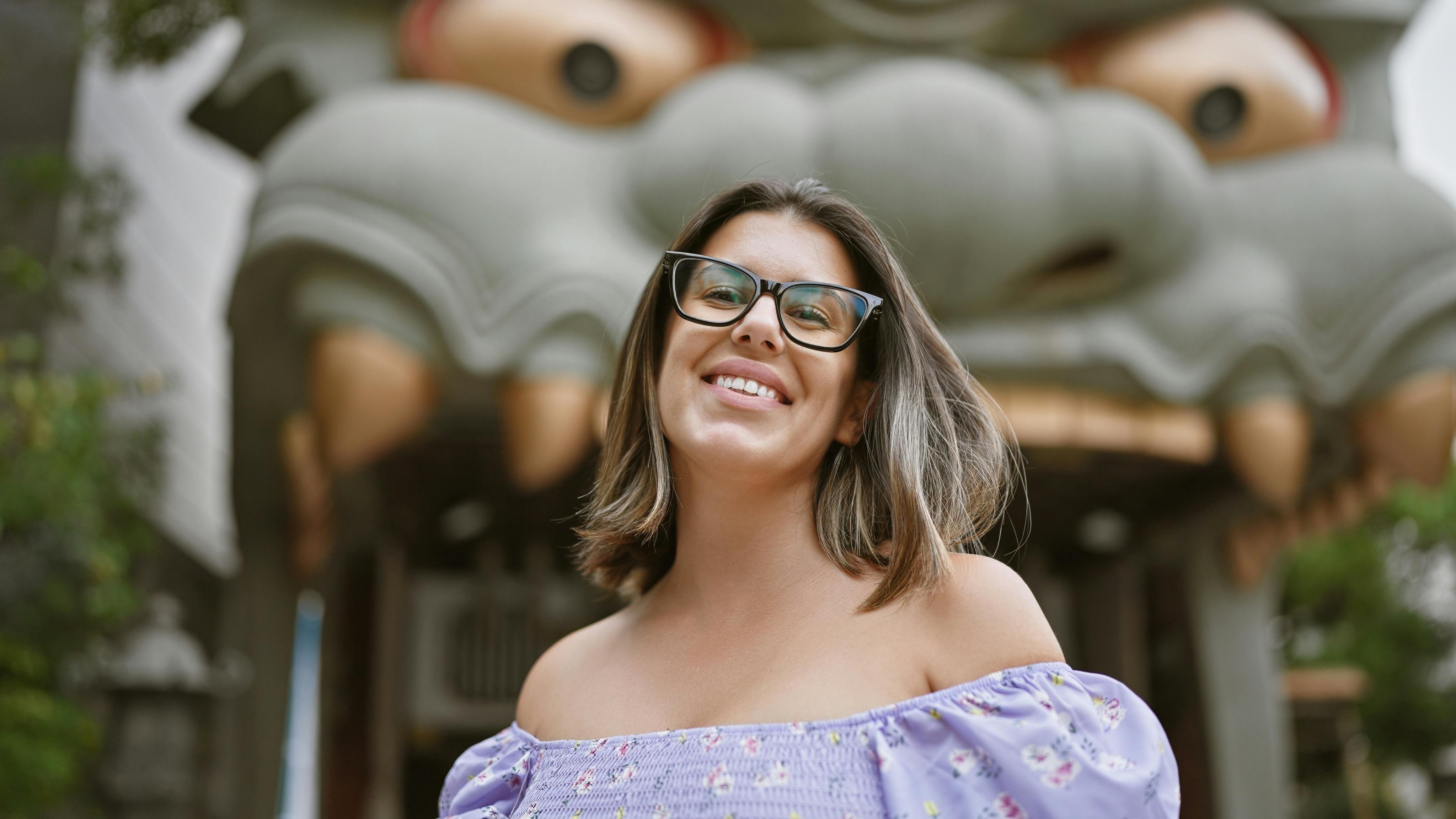 A smiling woman wearing glasses and a light purple off-the-shoulder top stands in front of a large, decorative dragon or lion head entrance, likely at an outdoor cultural site.