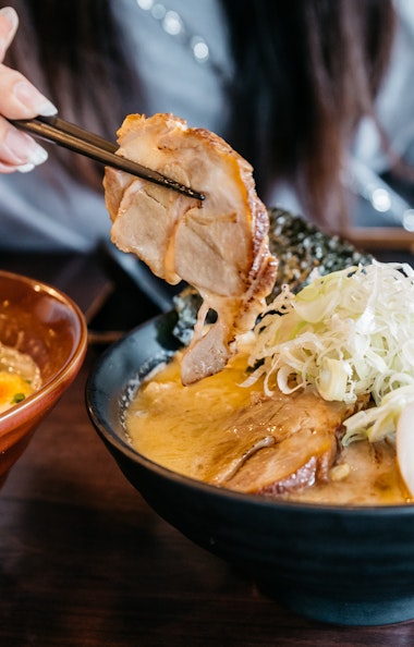 Hakata Ramen A person holds a slice of pork with chopsticks over a bowl of ramen, topped with sliced green onions and egg halves. Another bowl of ramen with green onions and eggs sits nearby on a wooden table.