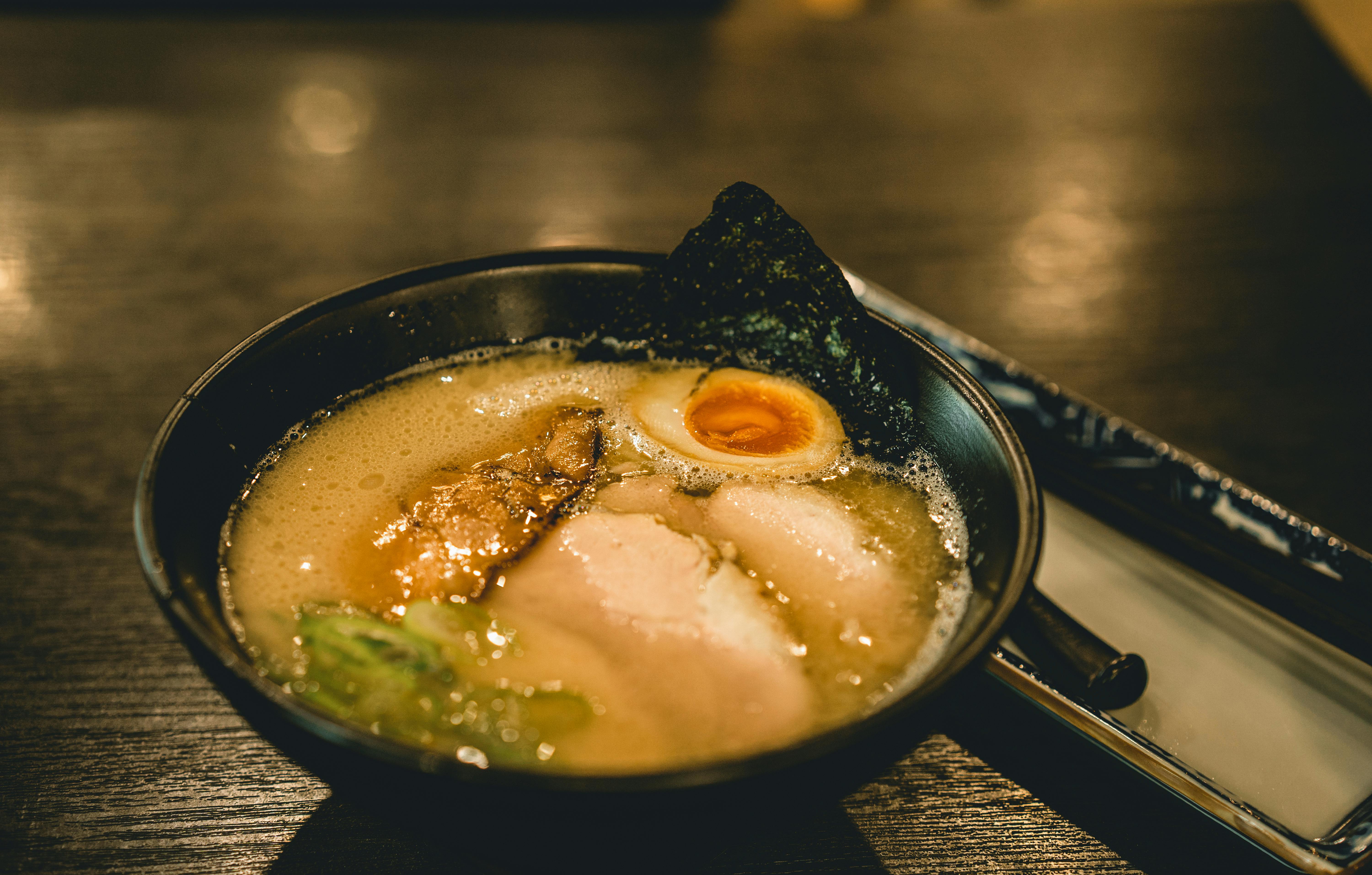 A bowl of ramen with broth, sliced pork, half a soft-boiled egg, green onions, and a sheet of seaweed, served on a dark wooden table with chopsticks beside the bowl.