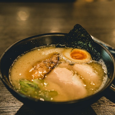 Hakata Ramen A bowl of ramen with broth, sliced pork, half a soft-boiled egg, green onions, and a sheet of seaweed, served on a dark wooden table with chopsticks beside the bowl.