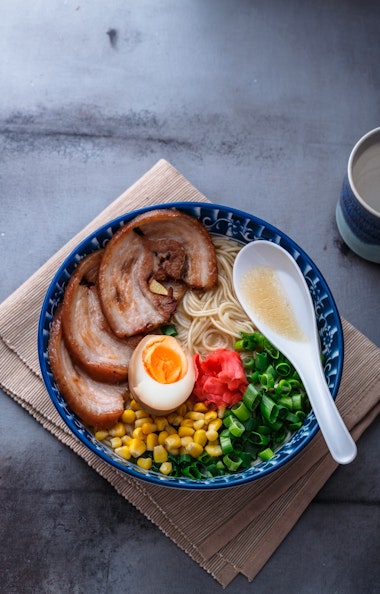 Hakata Ramen A bowl of ramen with sliced pork, half a soft-boiled egg, corn, green onions, pickled ginger, and noodles in broth, served with a white spoon on a blue patterned bowl, sitting on a bamboo mat.