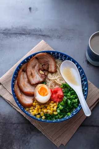 A bowl of ramen with sliced pork, half a soft-boiled egg, corn, green onions, pickled ginger, and noodles in broth, served with a white spoon on a blue patterned bowl, sitting on a bamboo mat.