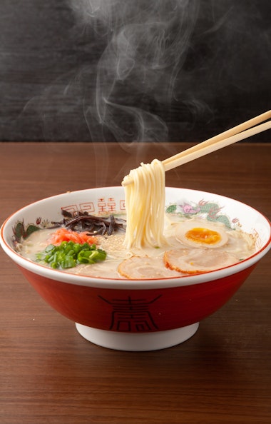 Hakata Ramen A steaming bowl of ramen with noodles lifted by chopsticks, topped with sliced pork, boiled egg halves, green onions, and mushrooms, served in a decorative bowl on a wooden table.
