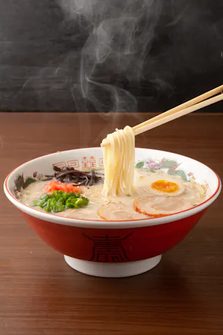 A steaming bowl of ramen with noodles lifted by chopsticks, topped with sliced pork, boiled egg halves, green onions, and mushrooms, served in a decorative bowl on a wooden table.