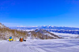 Two snowboarders sit on a snowy slope, facing a panoramic view of mountains and a clear blue sky, with snow-covered trees and a vast white landscape stretching into the distance.