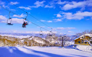 Skiers ride a chairlift over a snow-covered slope and trees on a sunny day, with a wooden lodge to the right and mountains visible in the distance under a bright blue sky with scattered clouds.