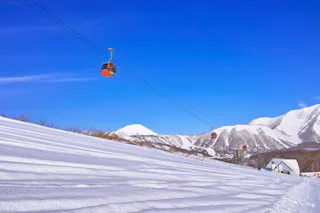 Red gondola cable cars travel above a snowy mountain slope under a clear blue sky, with snow-covered peaks in the background and a small building near the base.