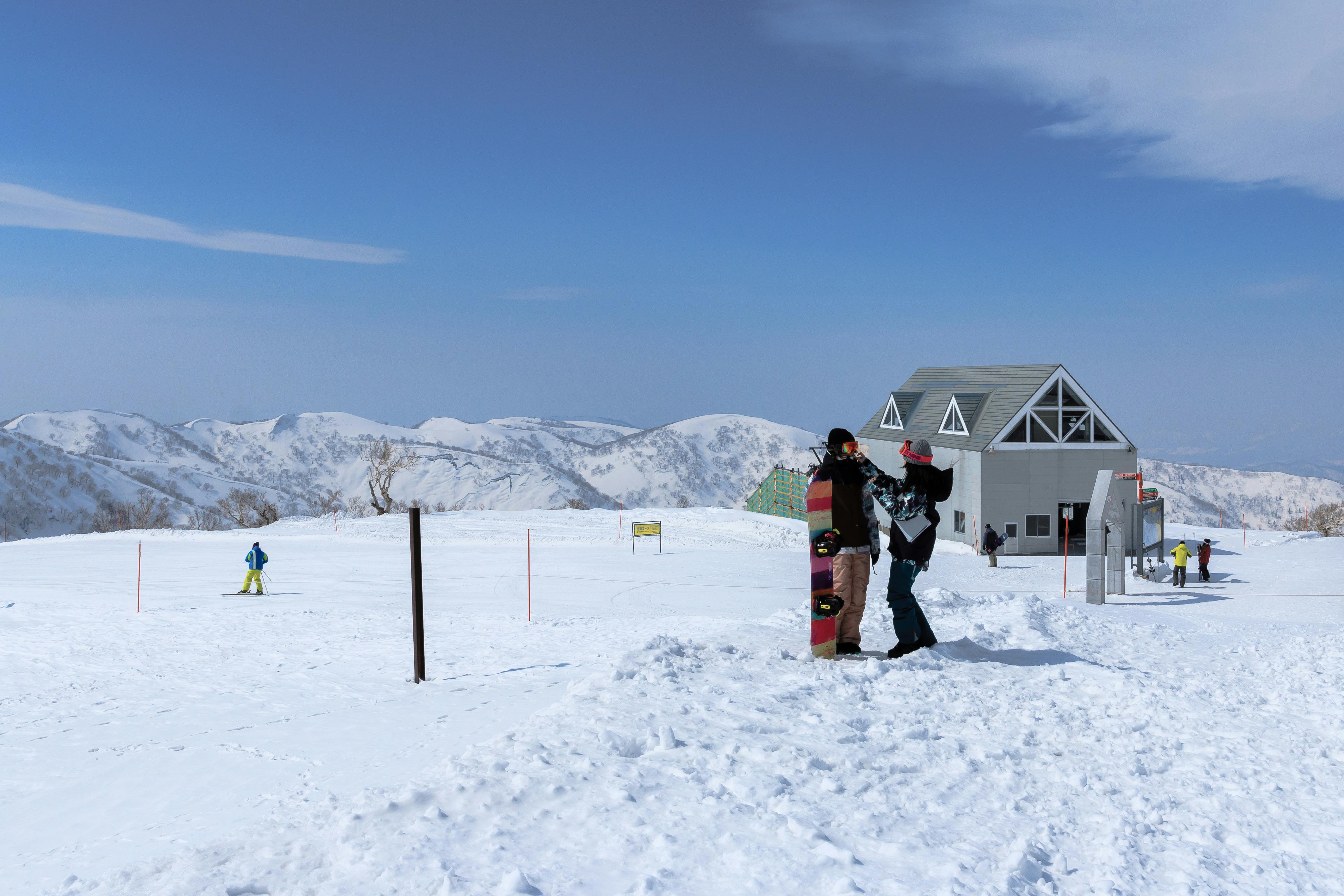 Snow-covered mountain scene with skiers and snowboarders near a small building. Snowboarders stand together in the foreground, while distant skiers move across the white landscape under a blue sky with mountains in the background.