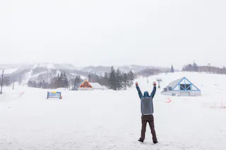 A person stands in the snow with arms raised, facing colorful A-frame buildings and snowy mountains in the background, under an overcast sky at a ski resort.
