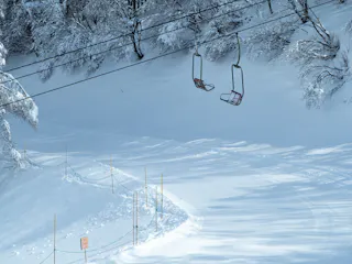 Empty ski lift chairs hang above a snow-covered slope lined with poles, netting, and a warning sign. Leafless trees coated in snow frame the scene, evoking a quiet, wintry atmosphere.
