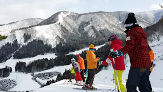 A group of skiers, including children and adults in colorful winter clothes, stand on a snowy slope with tall mountains and a parking lot visible in the background.