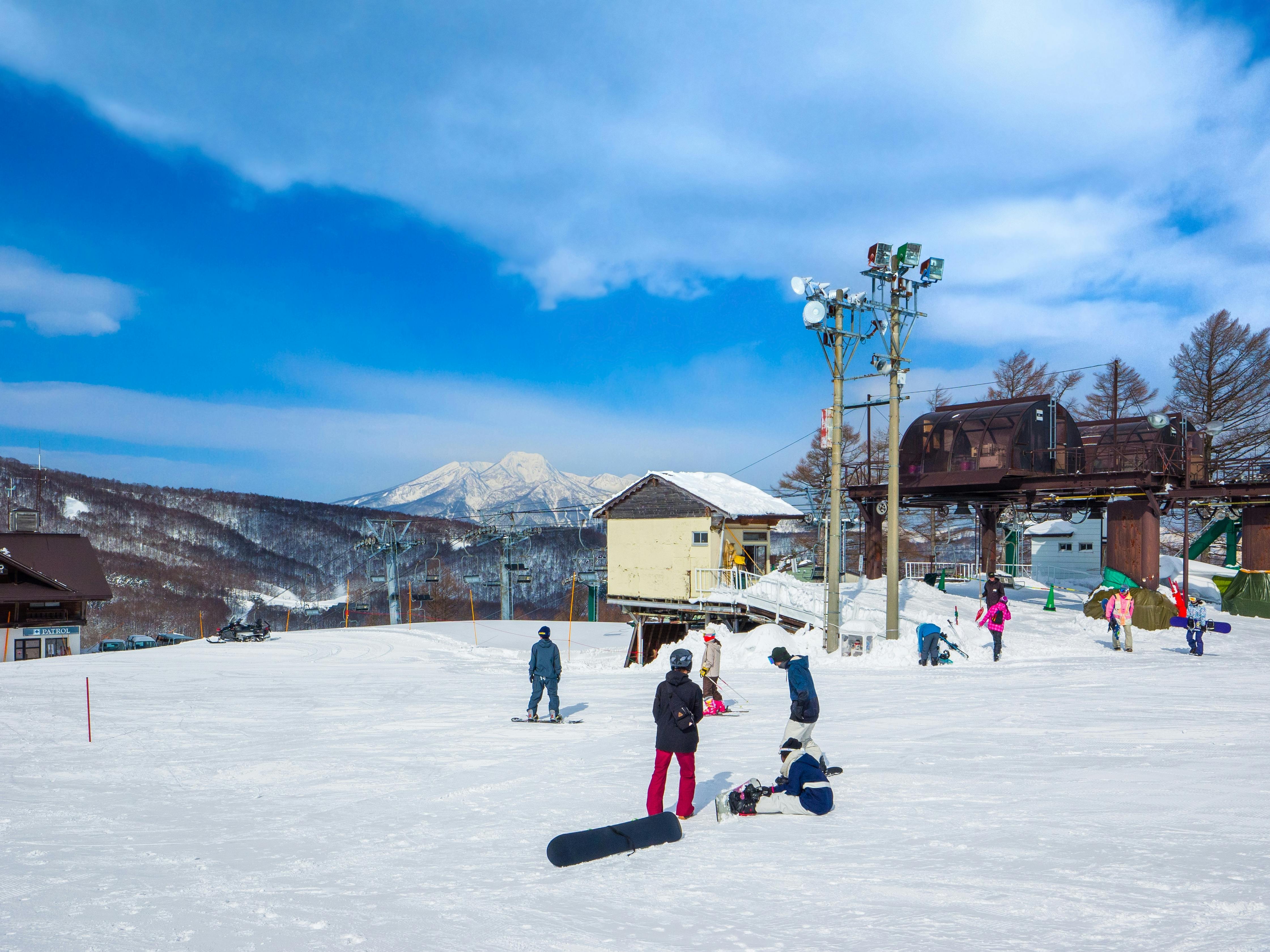 People skiing and snowboarding on a snowy mountain slope near ski lifts and buildings, with a clear blue sky and distant mountains in the background.