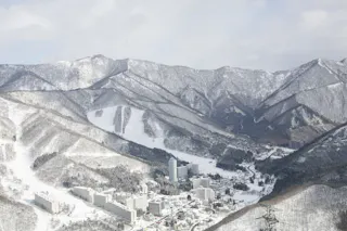 A winter landscape shows snow-covered mountains and a small town with tall buildings nestled in the valley below, surrounded by forested slopes and ski trails under a cloudy sky.