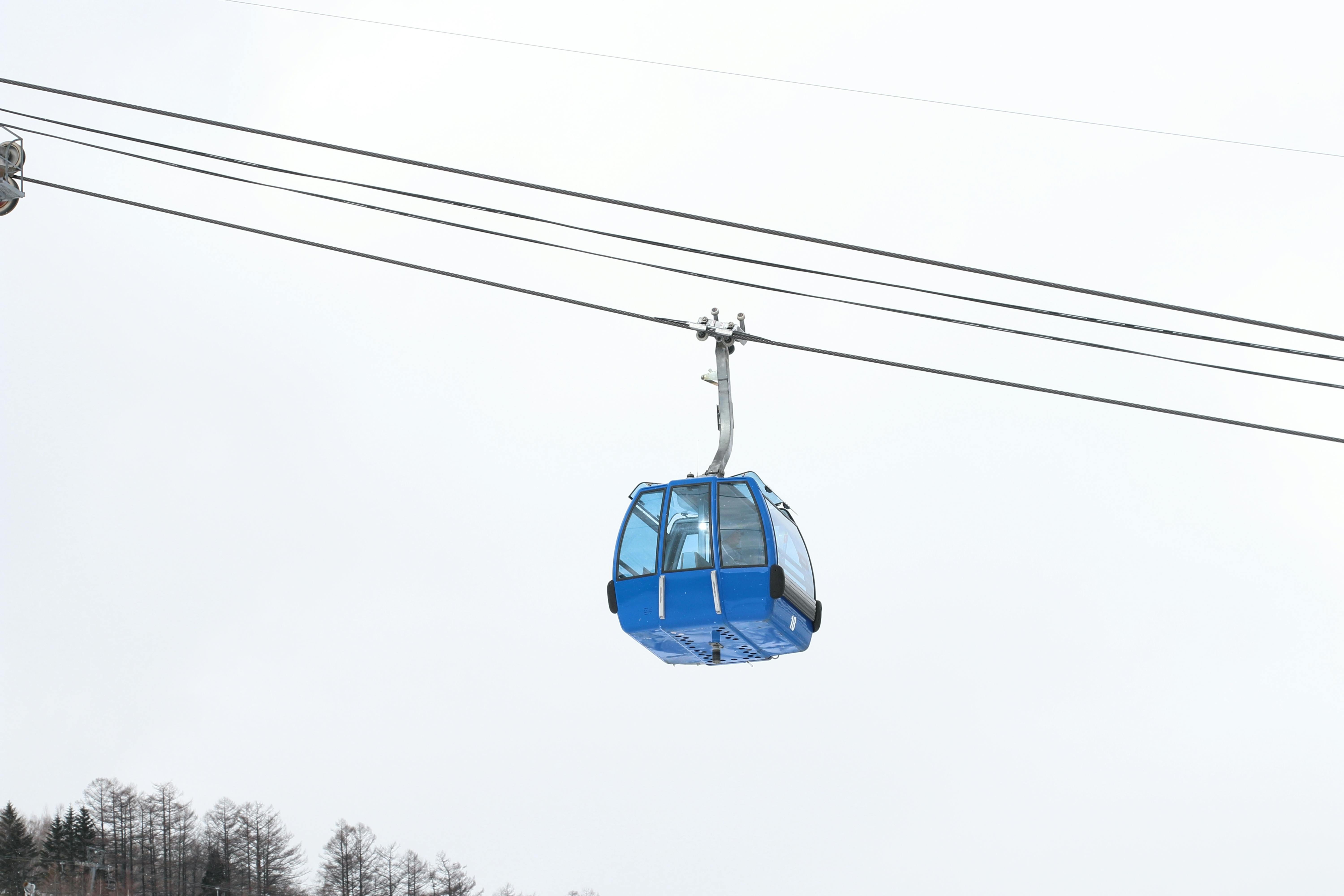 A blue gondola cable car travels along cables high above the ground, with a snowy, overcast sky in the background and tree tops visible below.