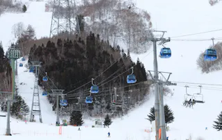 Blue ski gondolas and chairlifts move up a snowy mountain slope with scattered skiers below. Snow-covered trees and utility towers are visible in the background.