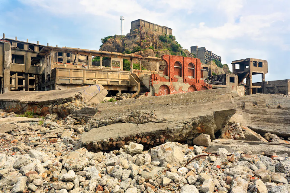 History of Nagasaki 버려진, 허물어져 가는 산업 단지로, 앞에는 부서진 콘크리트 구조물과 잔해가 있다. 배경에는 더 많이 낡은 건물들이 있는 언덕과 그 정상에 작은 등대가 구름 낀 하늘을 배경으로 있다.