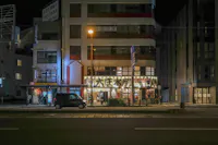 A city street at night with a lit storefront featuring Japanese signage. The building is illuminated, displaying various advertisements and lights. A car is parked on the street in front of the store, and a few people are visible near the entrance.