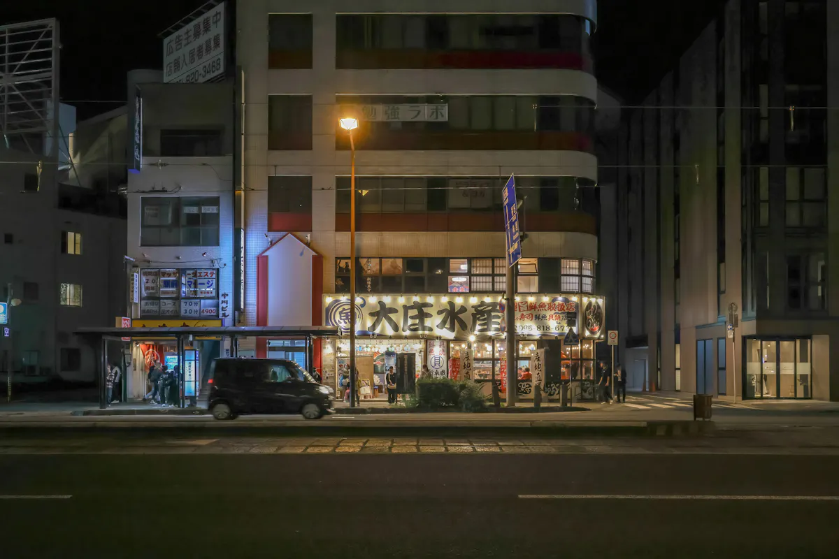 A city street at night with a lit storefront featuring Japanese signage. The building is illuminated, displaying various advertisements and lights. A car is parked on the street in front of the store, and a few people are visible near the entrance.