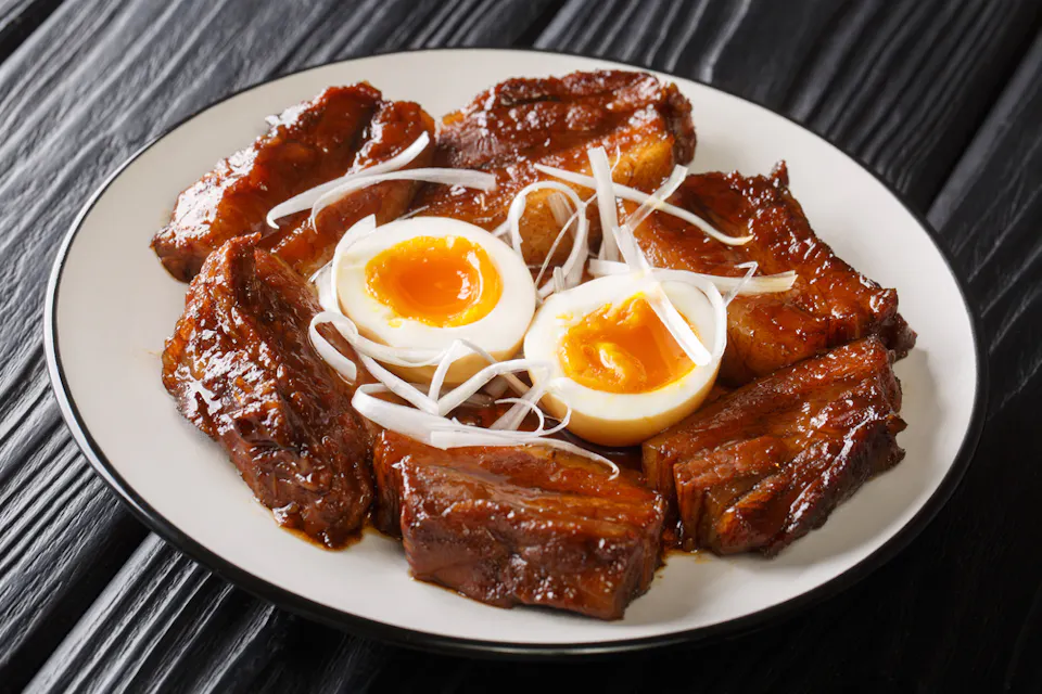 薄切りのキャラメル化された豚バラ肉のプレートに、細く切った白玉ねぎが添えられ、半分に切った卵の黄身が流れる柔らかい卵が2つ盛り付けられ、全てがダークウッドのテーブルにセットされている。