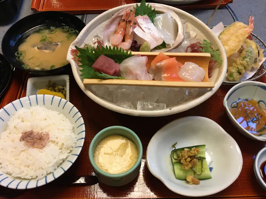 氷の上に刺身のボウル、蒸し白米、味噌汁、天ぷら、茶碗蒸し（蒸し卵のカスタード）、漬物、小さなきゅうりのサラダ、そしてディップソースを特徴とする日本の食事の広がり。