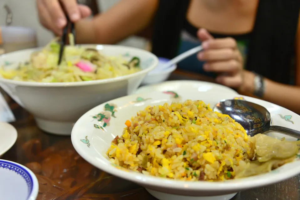 色とりどりのチャーハンの皿と大きな麺スープのボウルがあるテーブルセッティングのクローズアップ。人物が箸を持ち、麺のボウルから食べている。背景には木製の家具がある屋内のダイニングセッティングが示唆されている。