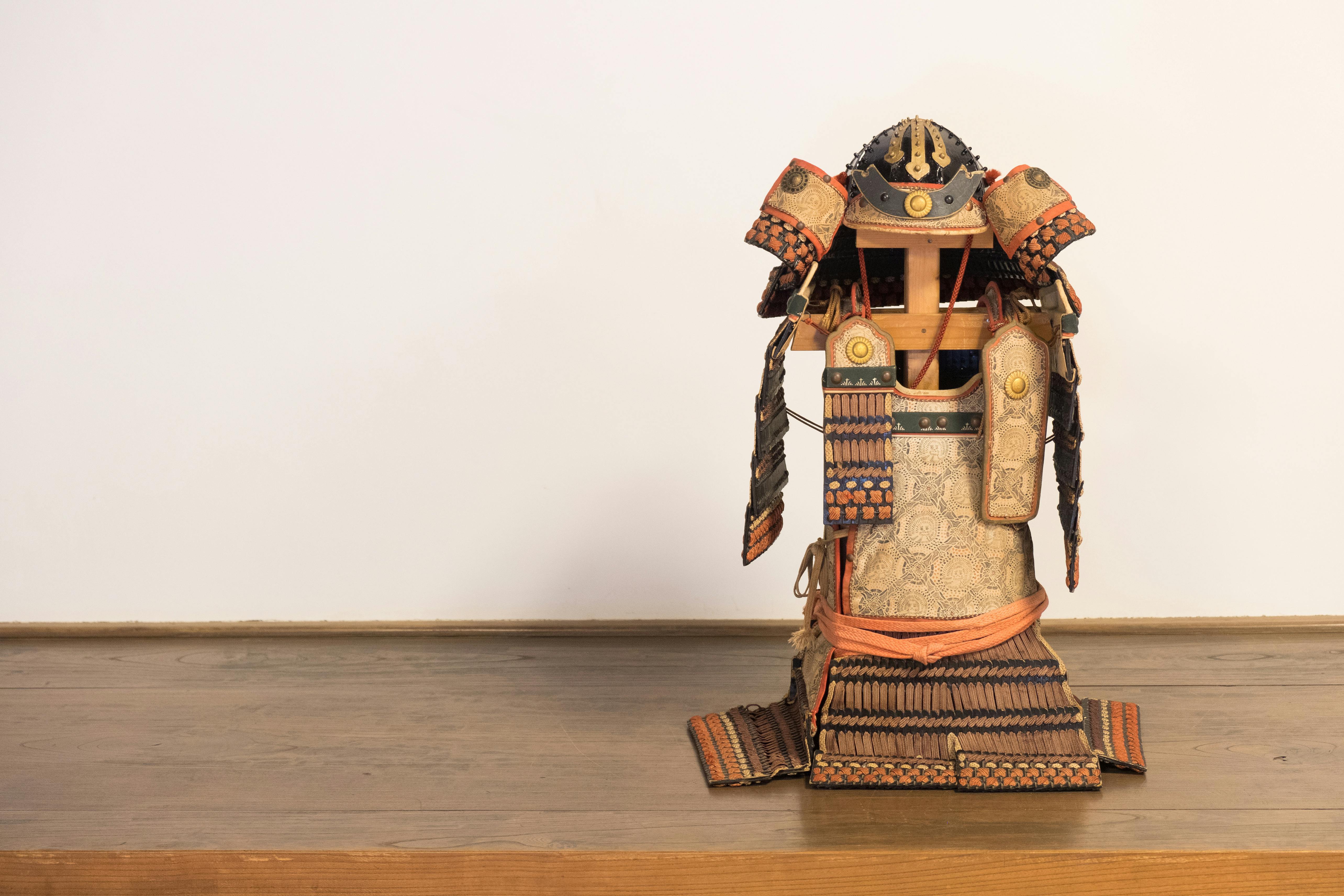 A traditional Japanese samurai armor set is displayed upright on a wooden surface against a plain, light-colored wall. The armor features intricate detailing and patterns with orange and dark accents.
