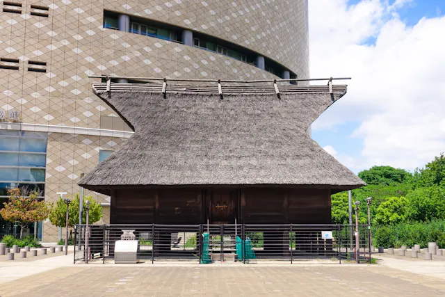 A traditional Japanese building with a thick thatched roof stands in front of a modern, patterned high-rise. The structure is surrounded by a metal fence, with greenery and a partly cloudy sky in the background.