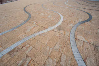 Wavy metallic strips embedded in a large expanse of textured, tan stone tiles create an abstract, modern pattern on the ground. The scene appears to be an urban plaza or open public space.