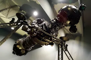 A large, metallic, intricate space probe or satellite model displayed indoors, featuring multiple antennas, domes, and structural supports, illuminated by overhead lighting.