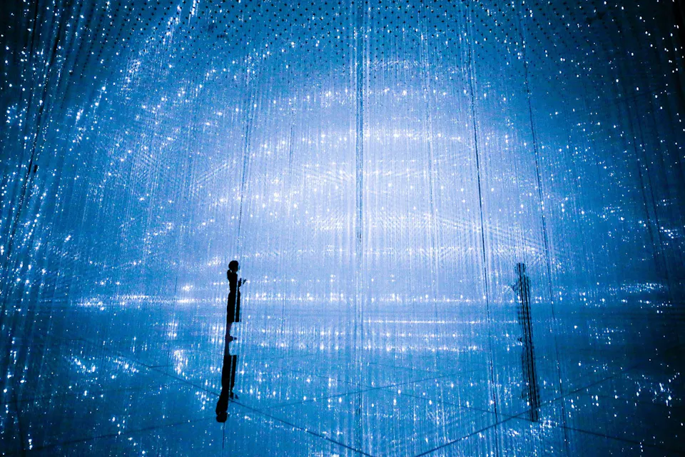A dimly lit room features ethereal blue lighting with delicate hanging strings or lights creating a starry effect. Silhouettes of two people are visible, adding to the mystical atmosphere. The scene has a futuristic, dreamlike quality.