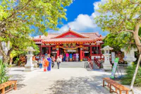A group of people, some in traditional clothing, stand in front of a vibrant red and white Japanese shrine surrounded by trees under a blue sky with scattered clouds.