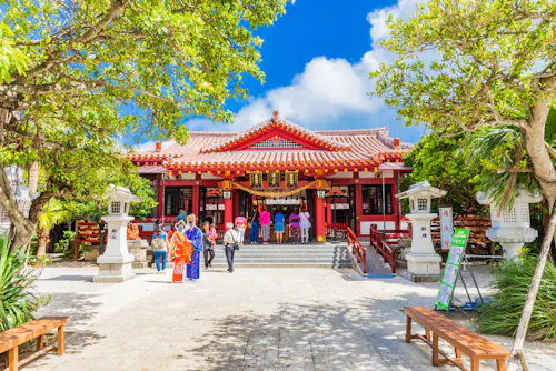 A group of people, some in traditional clothing, stand in front of a vibrant red and white Japanese shrine surrounded by trees under a blue sky with scattered clouds.